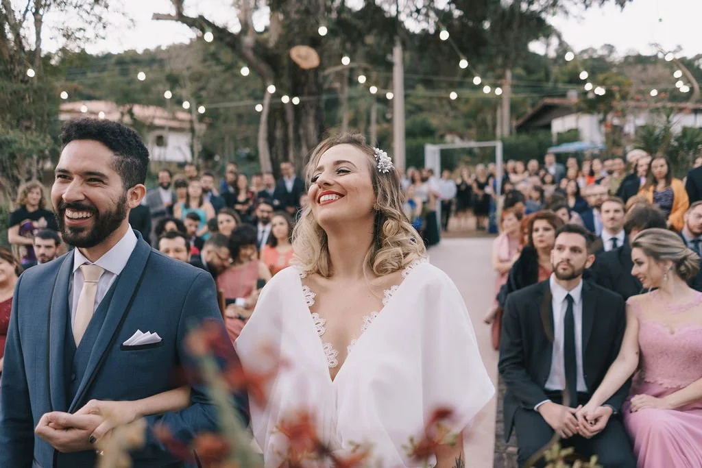 Casal de noivos sorrindo durante cerimônia de casamento ao ar livre com convidados ao fundo e decorações de luzes suspensas.