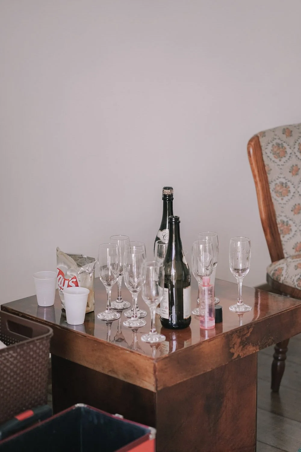 Mesa de madeira com taças de vidro, duas garrafas de bebida, um pacote de salgadinhos Lay's, copos descartáveis e um spray de desinfetante. Ao fundo, uma parede lisa e uma poltrona de estofado floral.