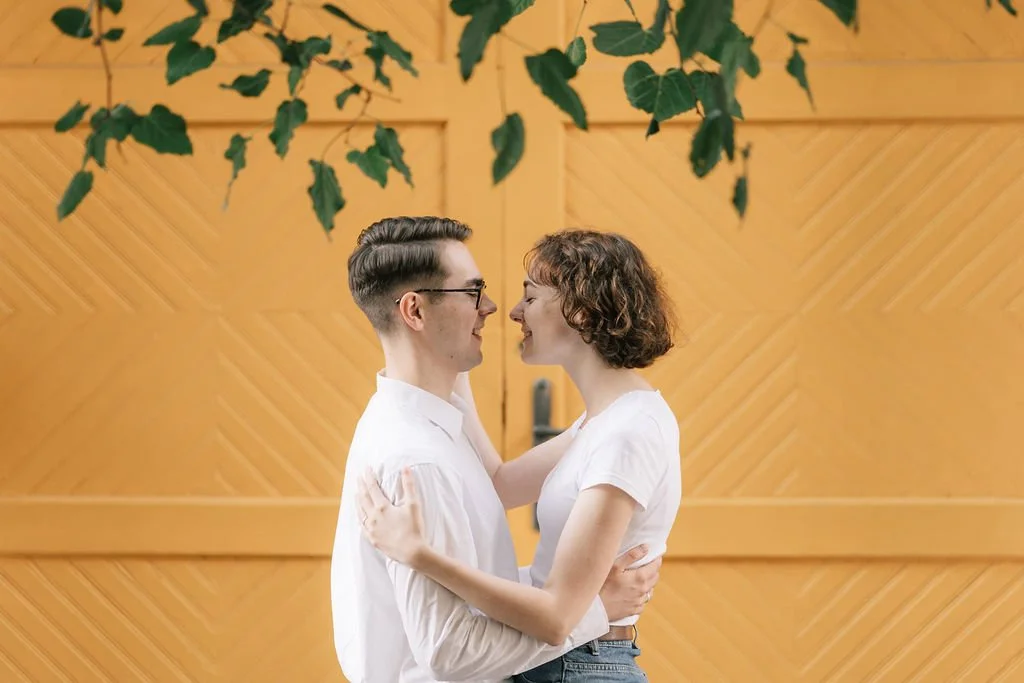 Casal sorrindo e se abraçando, olho nos olhos dentro de uma casa com paredes de madeira e folhas verdes penduradas no teto.