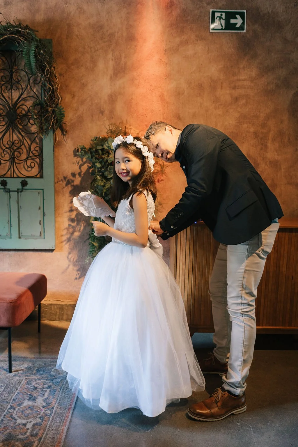 Menino ajustando o vestido de uma menina em um vestido de festa branca, enquanto ela sorri para a câmera.
