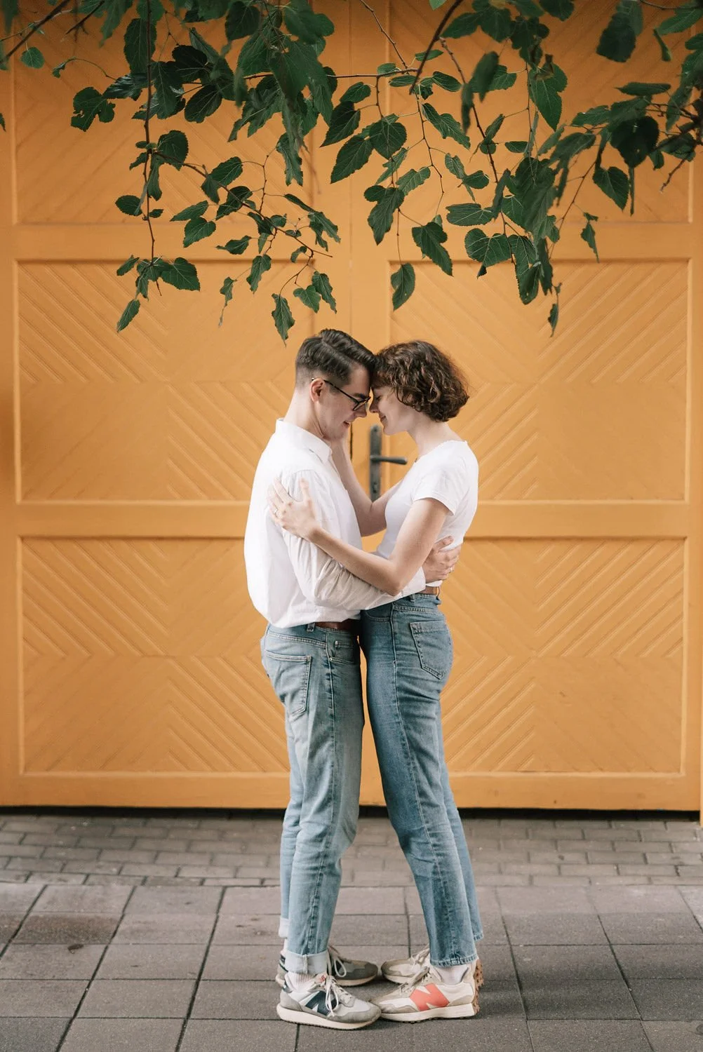 Casal jovem se abraçando, com cabeça junta, diante de uma porta de madeira e folhas verdes acima.