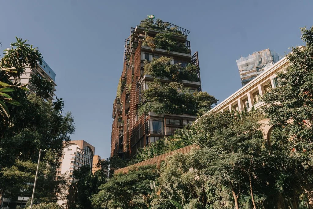 Prédio moderno com muitas plantas na fachada, cercado por árvores, em uma área urbana sob céu claro.