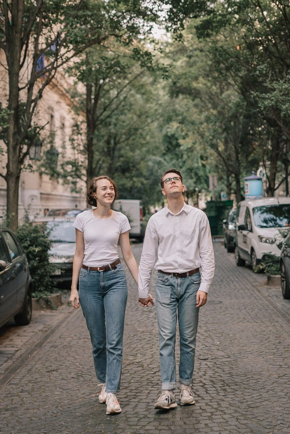 Casal andando de mãos dadas em uma rua arborizada, sorrindo e olhando para o céu.
