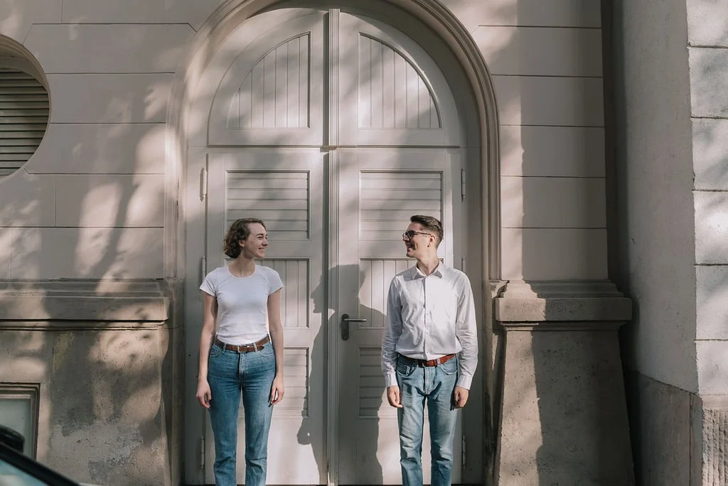 Homem e mulher sorrindo um para o outro na frente de uma porta grande e arcos, com sombra de árvore na parede