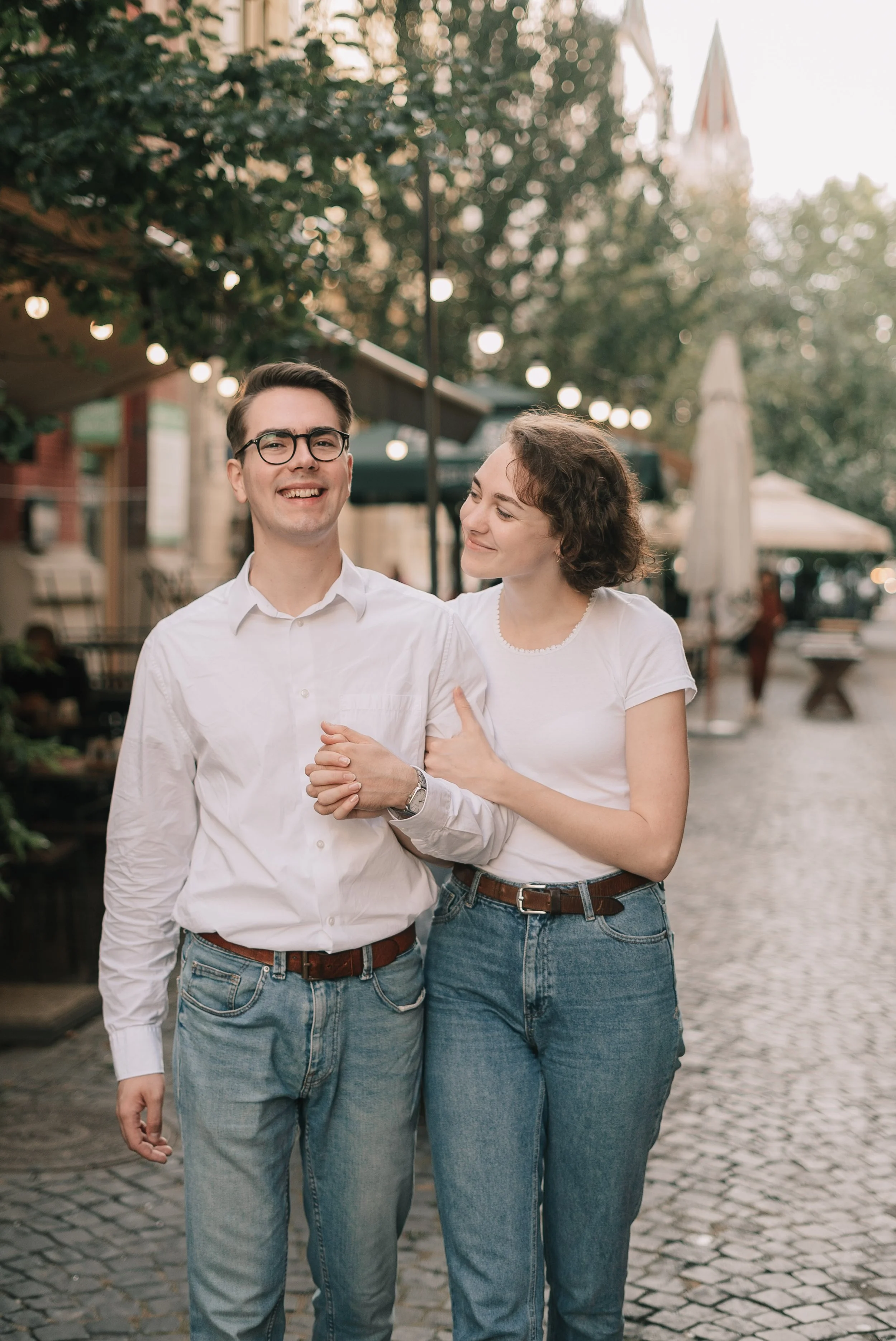Um casal andando de mãos dadas na rua, sorrindo e se olhando com carinho, em um ambiente ao ar livre com luzes decorativas e árvores ao fundo.