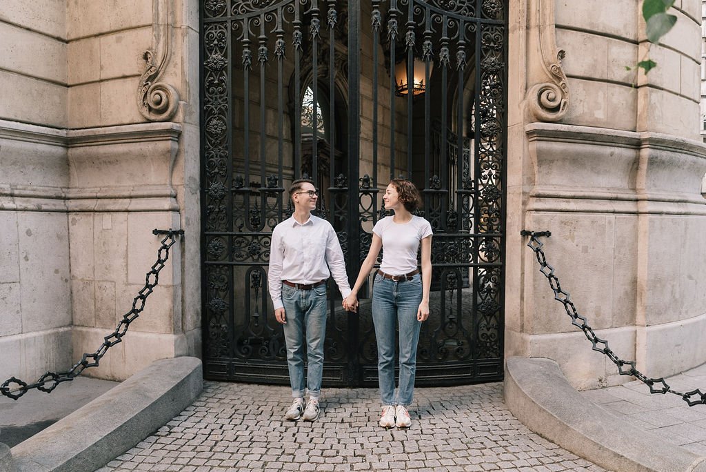Casal segurando as mãos em frente a uma porta de ferro decorativa em um edifício de pedra