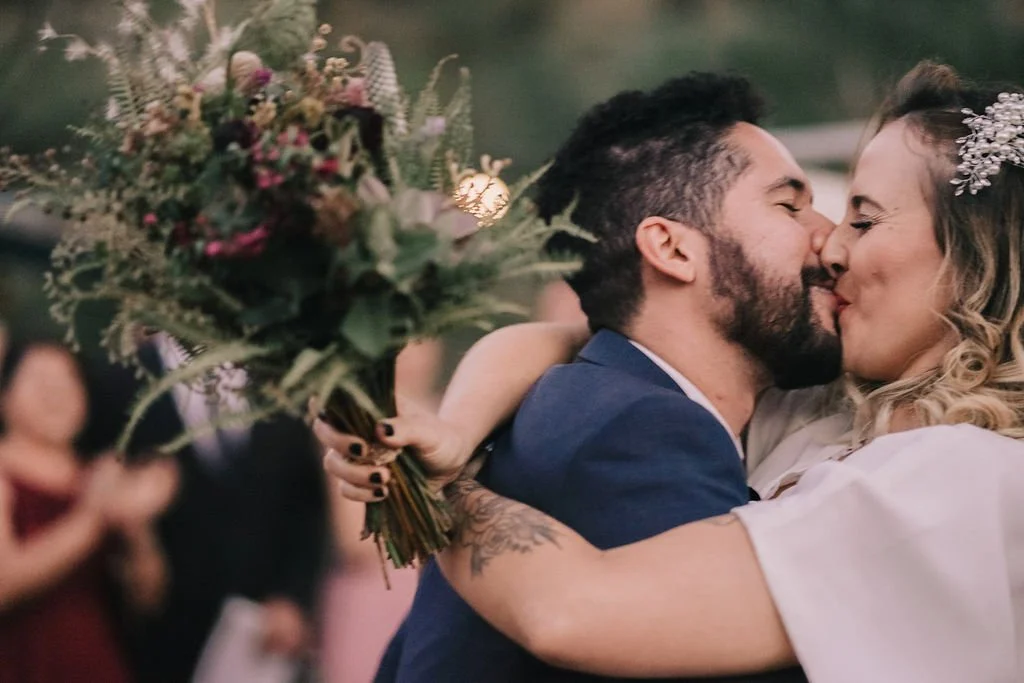 Casal se beijando, possivelmente no casamento, com uma mulher segurando um buquê de flores ao fundo.
