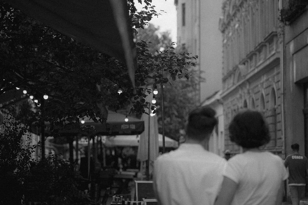 Casal caminhando na rua de noite, com iluminação decorativa e lojas ao fundo, em um ambiente urbano.