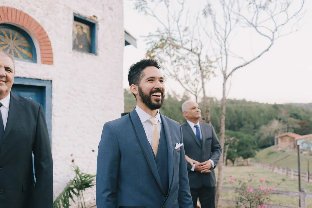Homem sorridente de terno azul e gravata, ao ar livre, em evento social, com duas outras pessoas ao fundo e natureza ao redor.