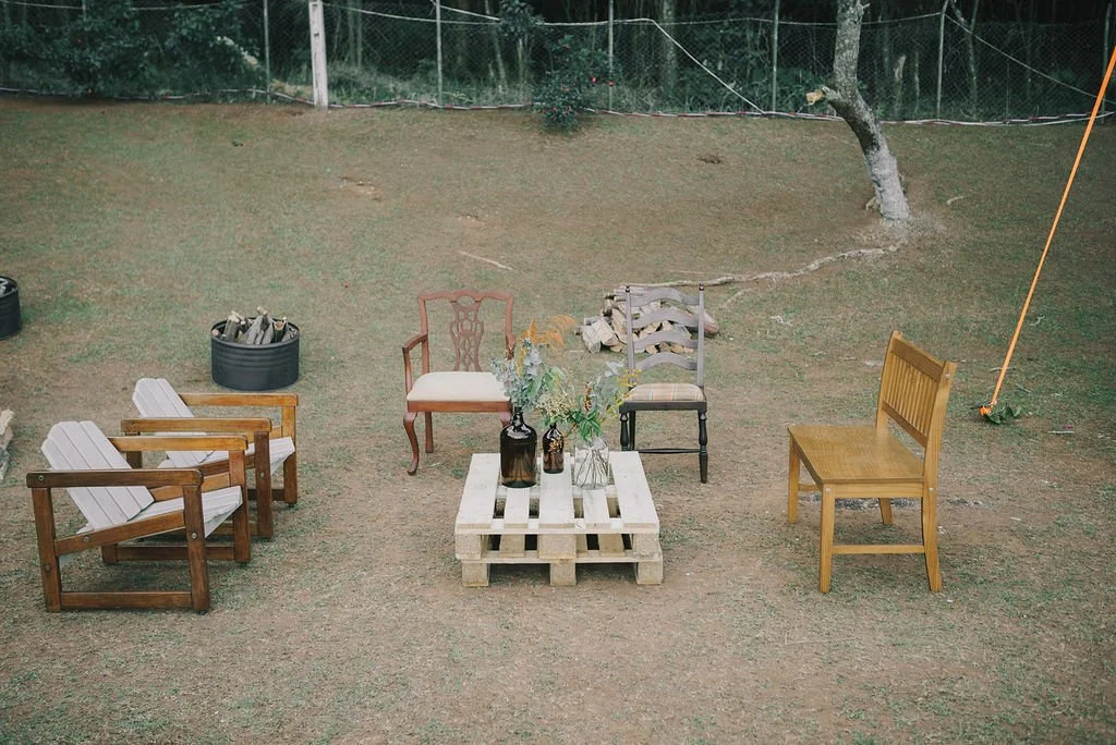 Móveis de jardim com cadeiras de madeira e paletes, uma mesa com vasos de plantas, em um ambiente ao ar livre com grama e uma árvore