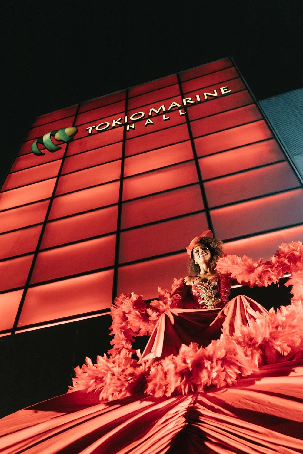 Mulher vestida com roupagem teatral vermelha e rosa, com penas, posando diante de um prédio iluminado com letreiro "Tokio Marine" à noite.