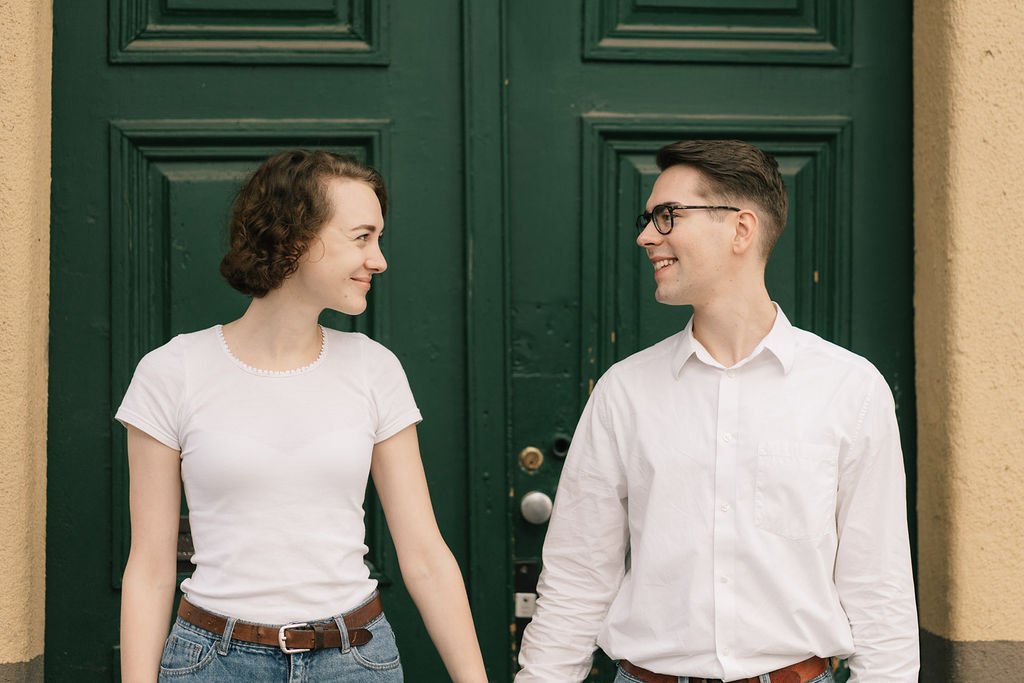 Um casal jovem sorrindo um para o outro segurando as mãos na frente de uma porta verde.