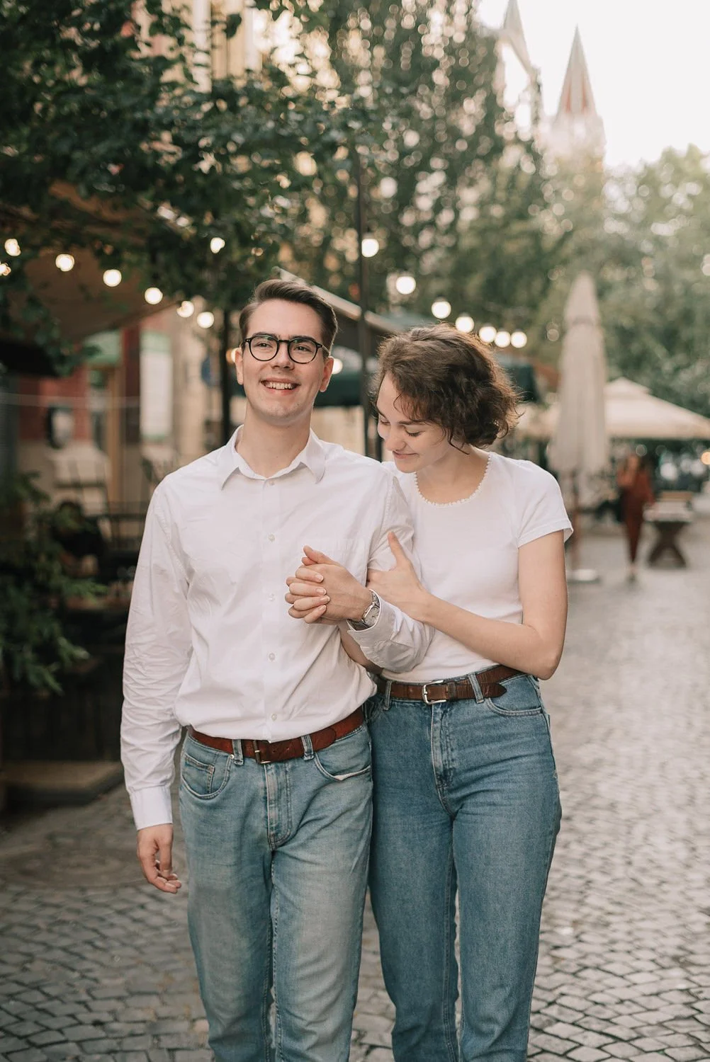 Casal feliz caminhando de mãos dadas pelo calçadão com árvores, luzes e edifícios ao fundo.
