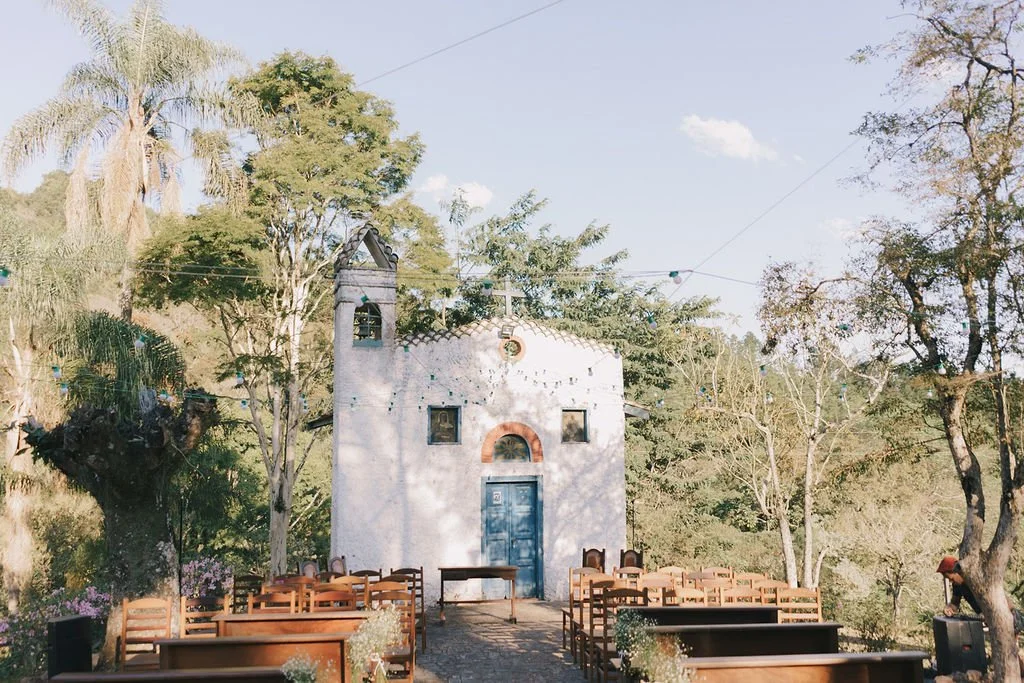 Pequena capela branca com portas azuis, rodeada por árvores e cadeiras de madeira, decorada com luzes suspensas, em um ambiente ao ar livre.