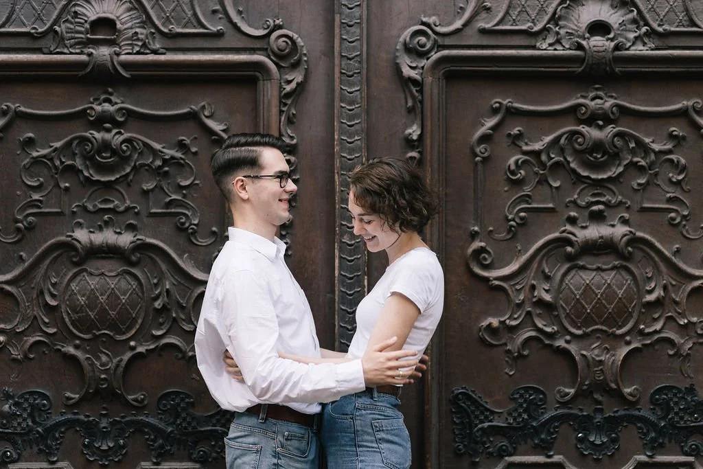 Um casal sorridente de mãos dadas em frente a uma porta de madeira com detalhes decorativos. Eles estão se olhando com carinho, usando roupas casuais brancas e jeans.