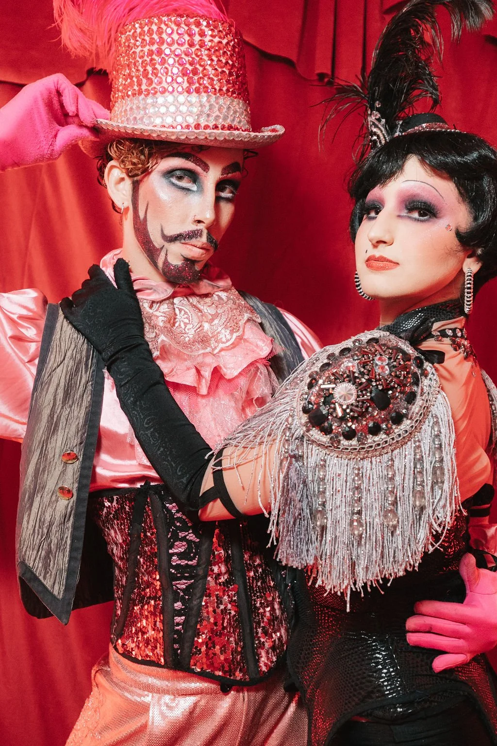Dois performers em trajes de shows de cabaré com figurinos elaborados e maquiagem dramática, posando de forma expressiva com fundo vermelho.