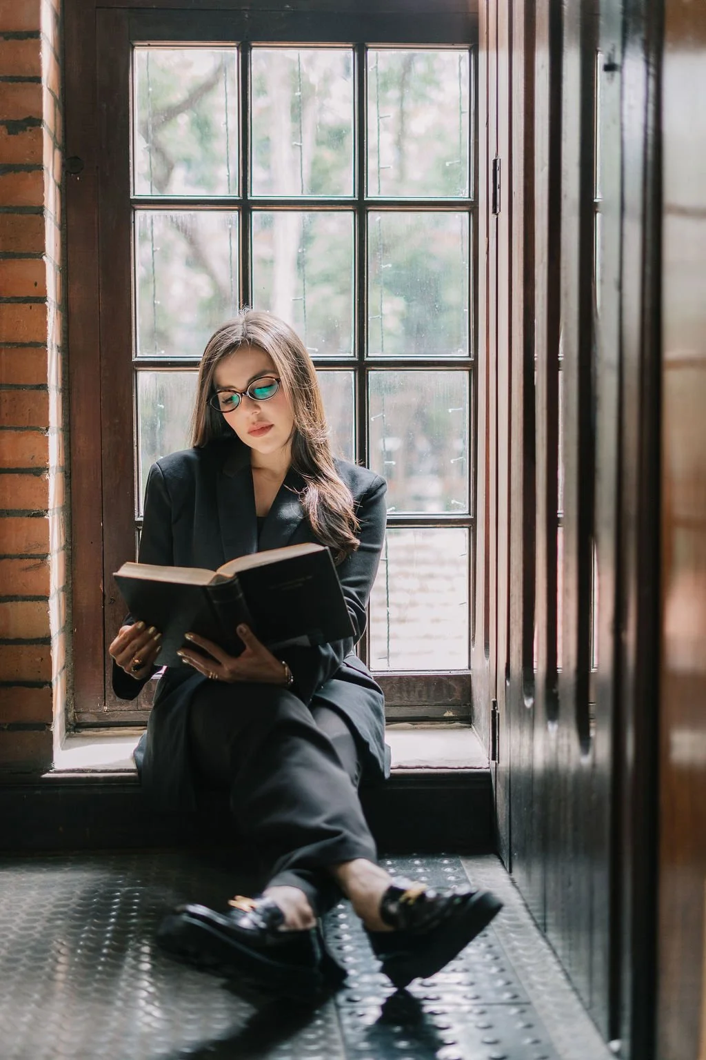 Jovem mulher de óculos escuros usando um blazer preto, sentado no degrau de uma janela de madeira, lendo um livro em um ambiente com paredes de tijolos. A iluminação natural entra pela janela.