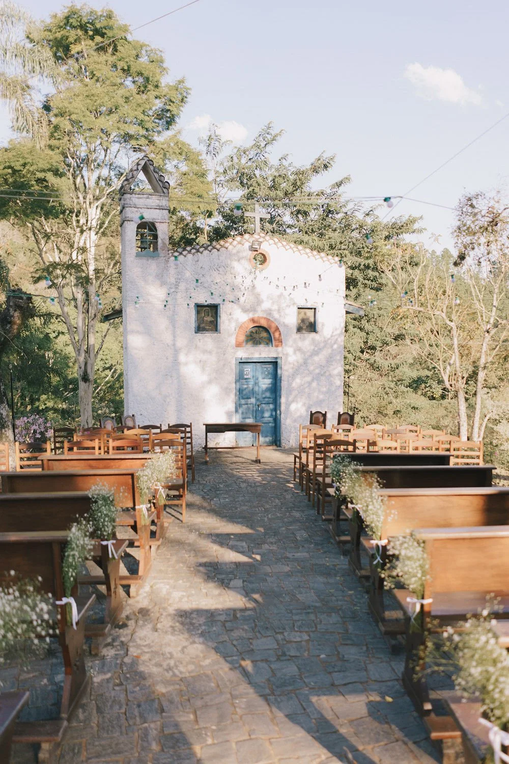 Cerimônia ao ar livre em frente a uma pequena igreja branca com porta azul e janelas pequenas, decorada com flores e luzes, em uma área aberta cercada por árvores.