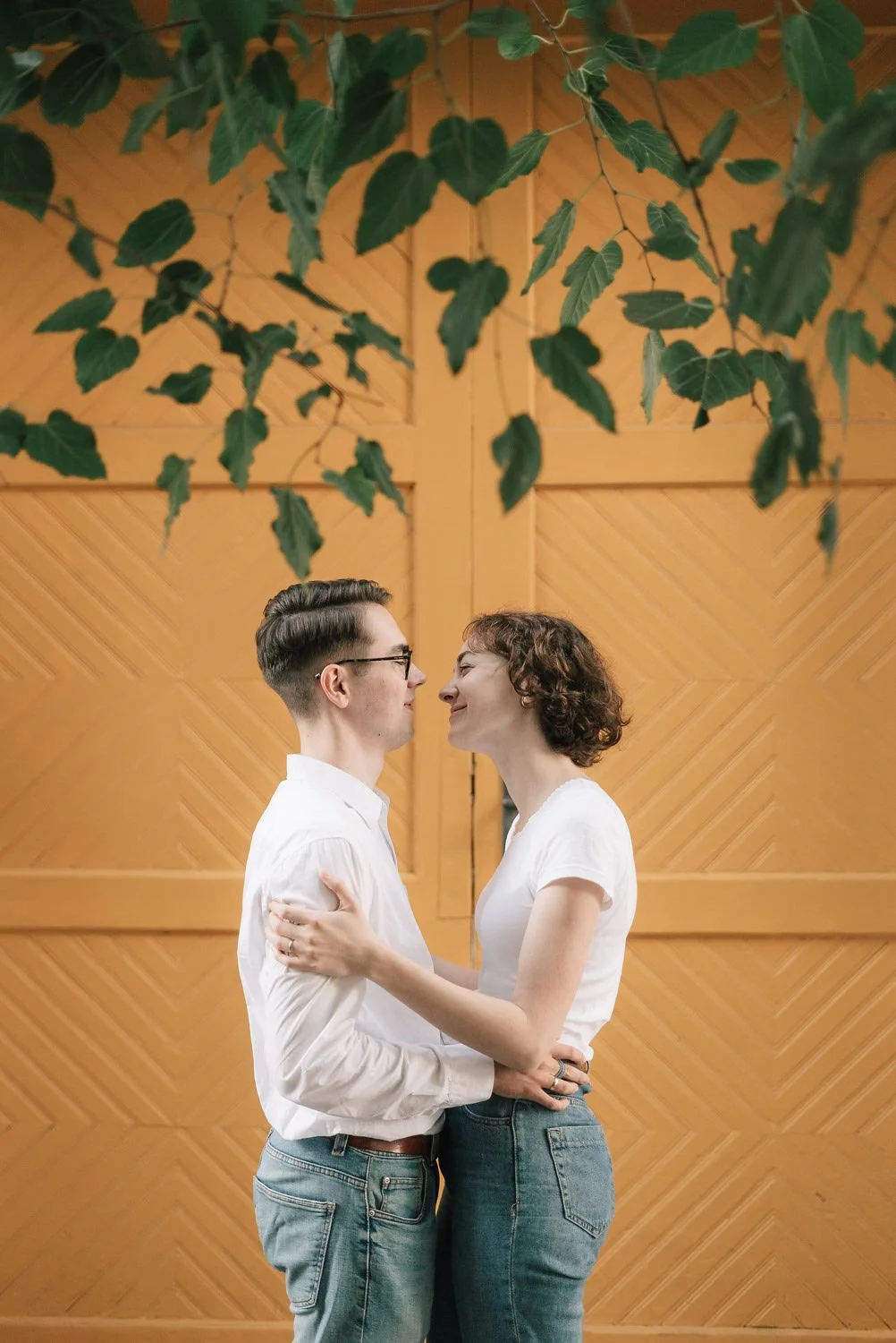 Casal adulto de frente, segurando um ao outro, sorrindo e quase se beijando, com fundo de parede de madeira e folhas verdes suspensas na parte superior.