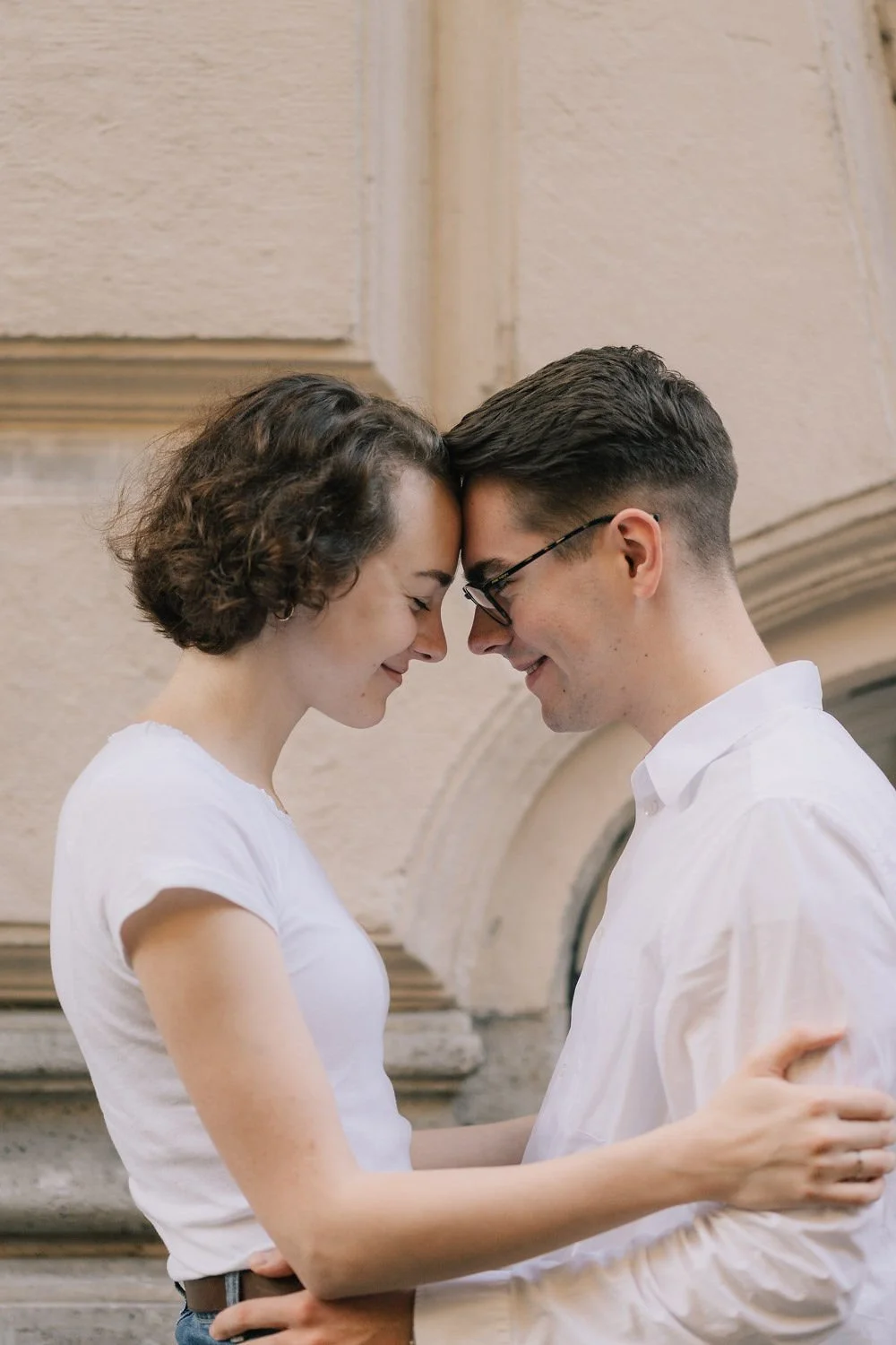 Um casal com testa e nariz tocando, sorrindo e abraçados, em um ambiente interno com parede de pedra ao fundo.