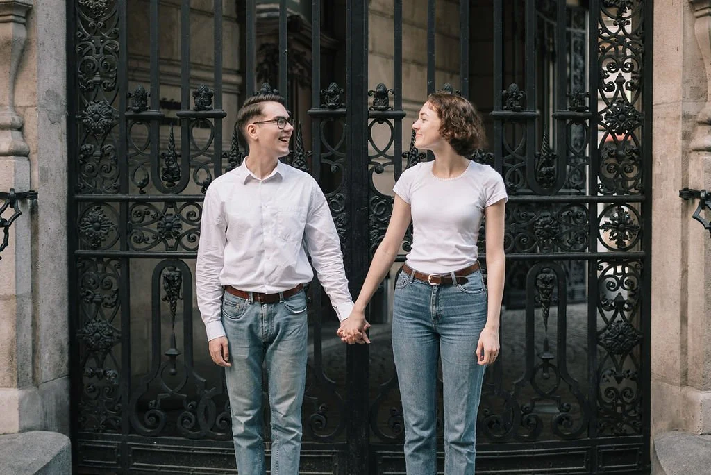 Duas pessoas segurando as mãos e sorrindo na frente de um portão de ferro decorado, ao ar livre.
