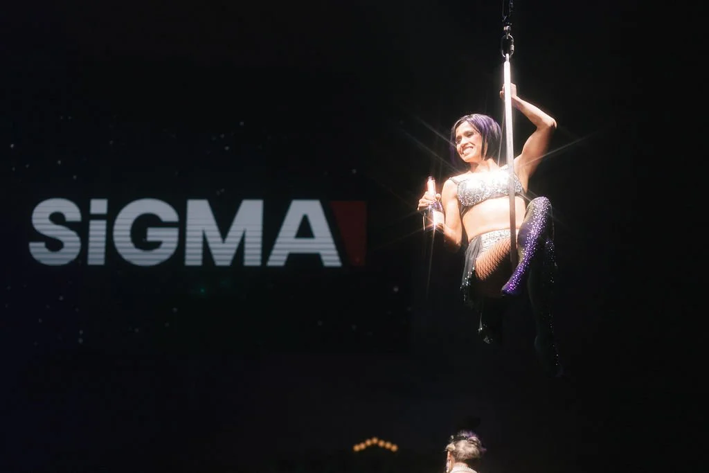 Mulher com vestido brilhante e meia arrastão, segurando uma garrafa de champanhe, naquilo que parece ser um palco ou evento, com a logo 'SIGMA' ao fundo.