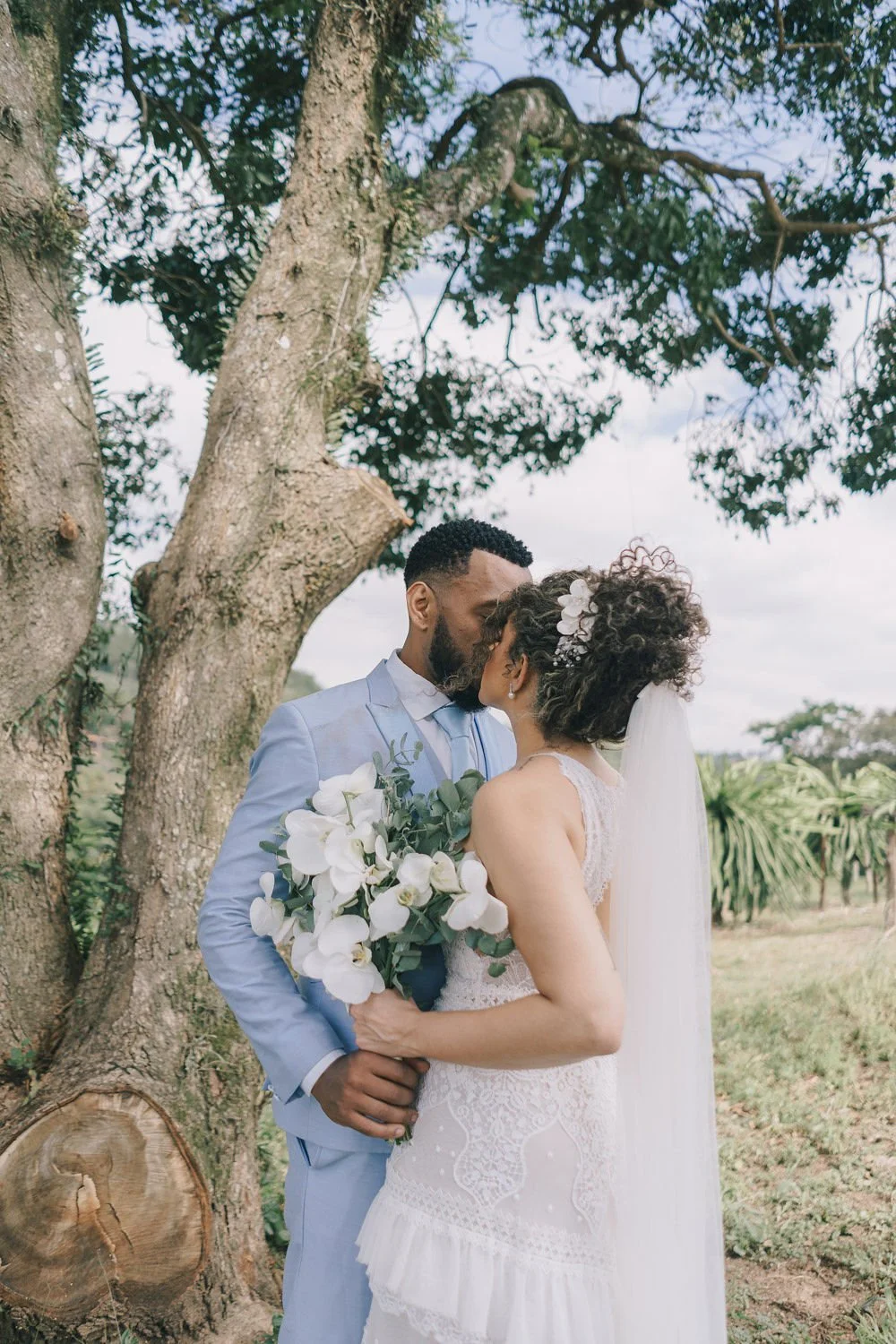 Casal de casamento se beijando ao ar livre, com árvores ao fundo, ela segurando um buquê de flores brancas.