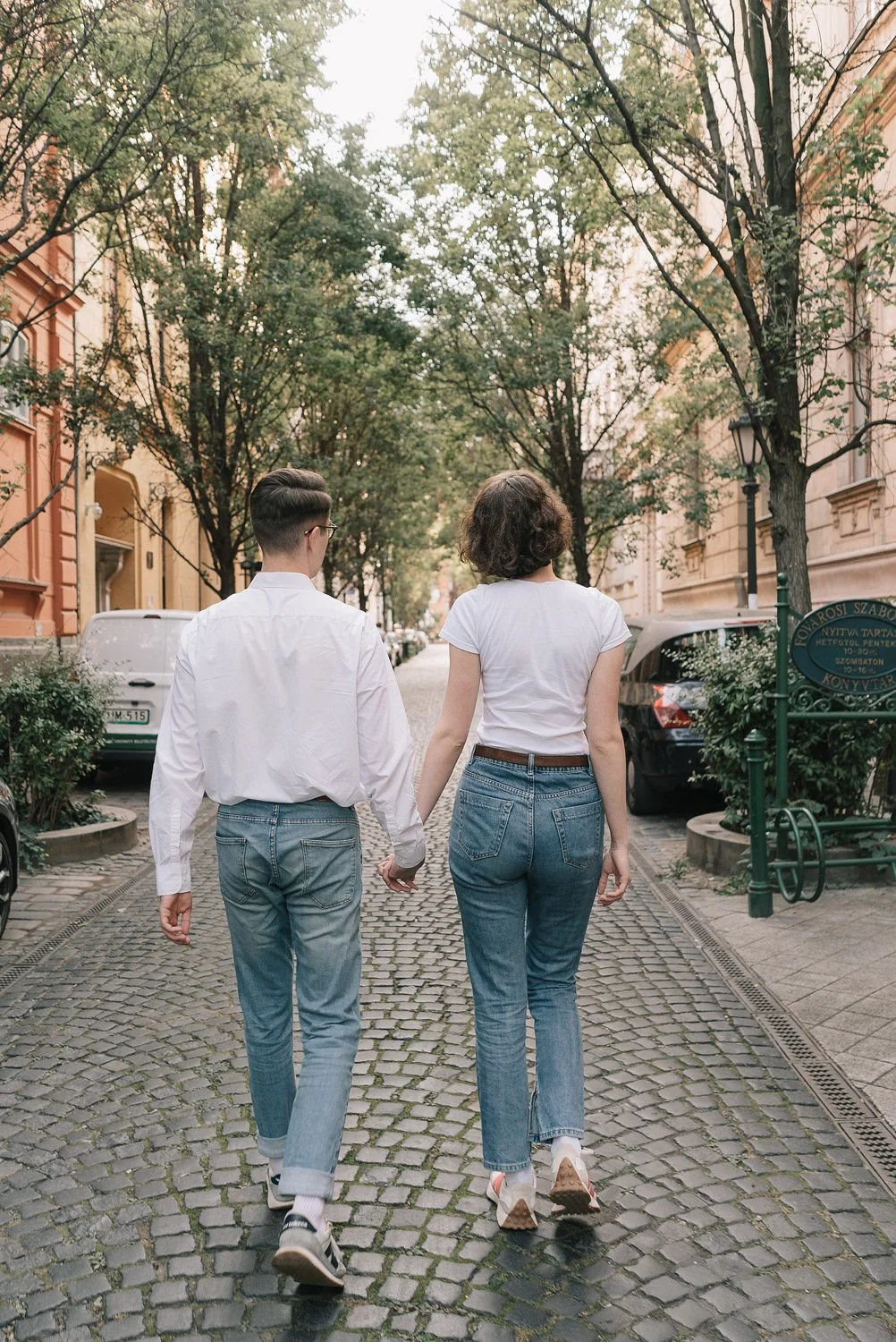 Casal caminhando de mãos dadas por uma rua de paralelepípedo com árvores e edifícios ao fundo, de costas, durante o dia.