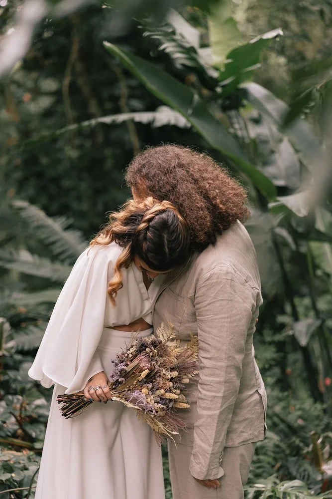 Casal de noivos com roupas claras trocando um beijo na testa, segurando um buquê de flores secas, em um ambiente com vegetação densa e folhagem.
