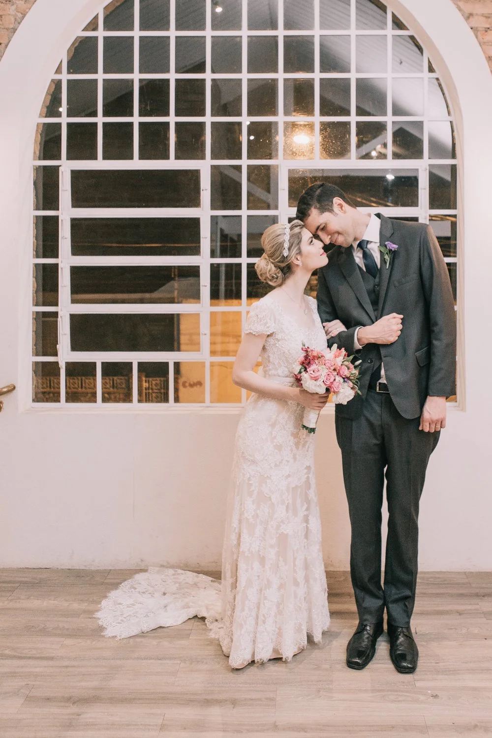 Casal de noivos em abraço íntimo durante casamento, ela segurando buquê de flores, no interior de uma sala com grande janela de vidro