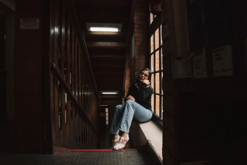 Mulher sentada na janela de uma escada de madeira, olhado para o lado, com iluminação natural vindo de uma janela grande ao lado.