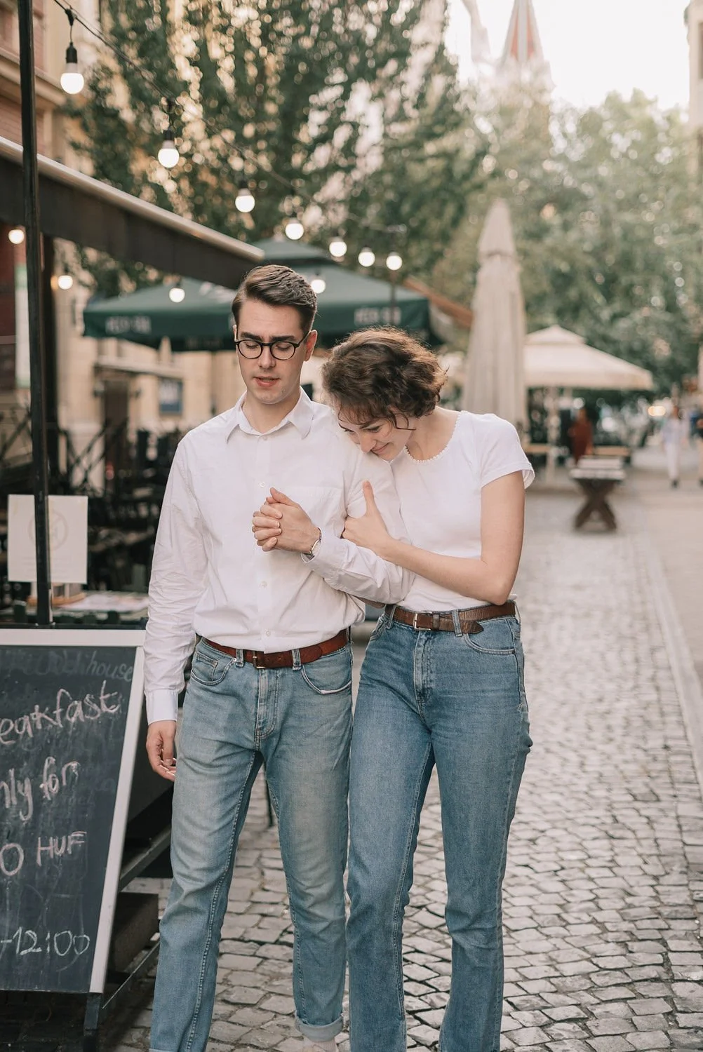 Casal de jovens caminhando de mãos dadas na rua, com iluminação de luzes decorativas e árvores em fundo, em um ambiente ao ar livre.