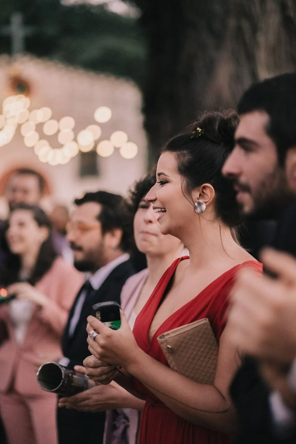 Mulher sorrindo em um evento social, usando vestido vermelho, com cabelo preso, segurando um telefone celular e um clutch
