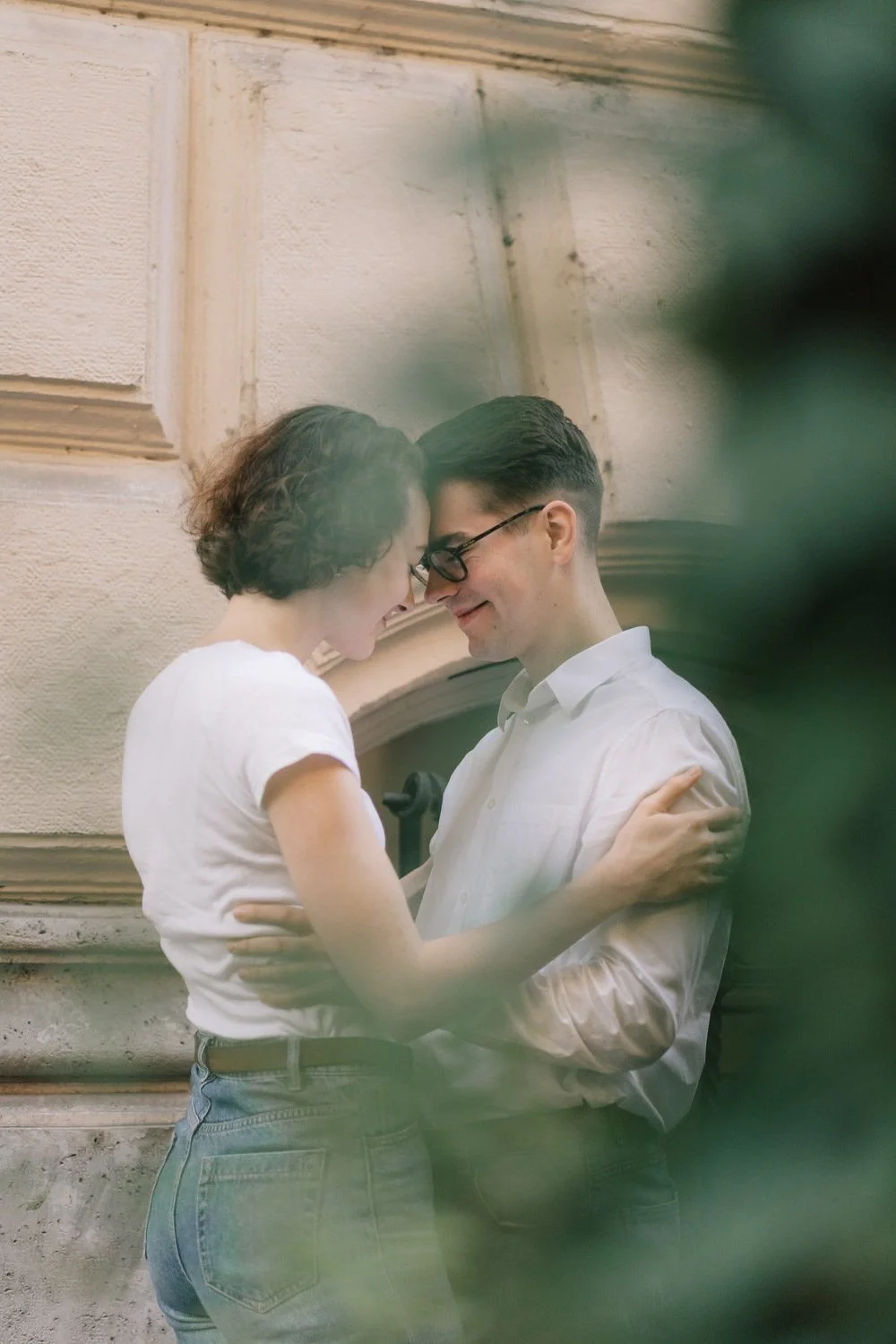 Um casal jovem, com roupas brancas, se abraçando e sorrindo, com uma parede de fundo em um ambiente interno, parcialmente encoberto por uma área verde borrosa na frente.