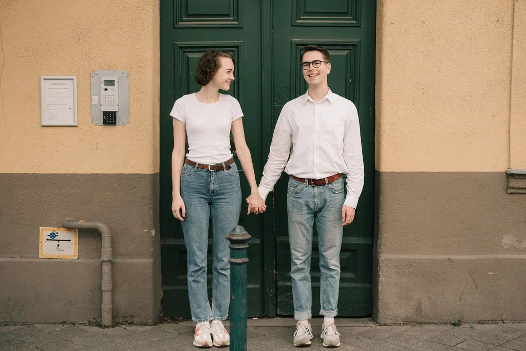 Um casal sorridente segurando as mãos em frente a uma porta verde, na rua.
