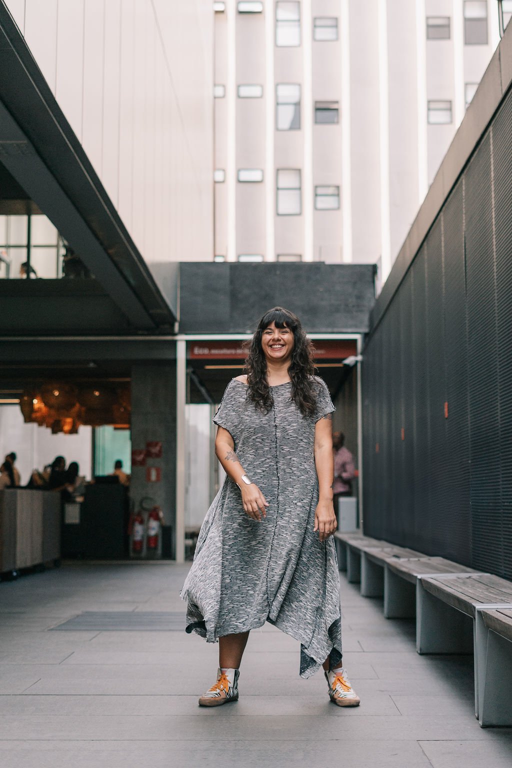 Mulher sorridente de vestido cinza com tênis laranja, usando um relógio, em um espaço urbano com arquitetura moderna