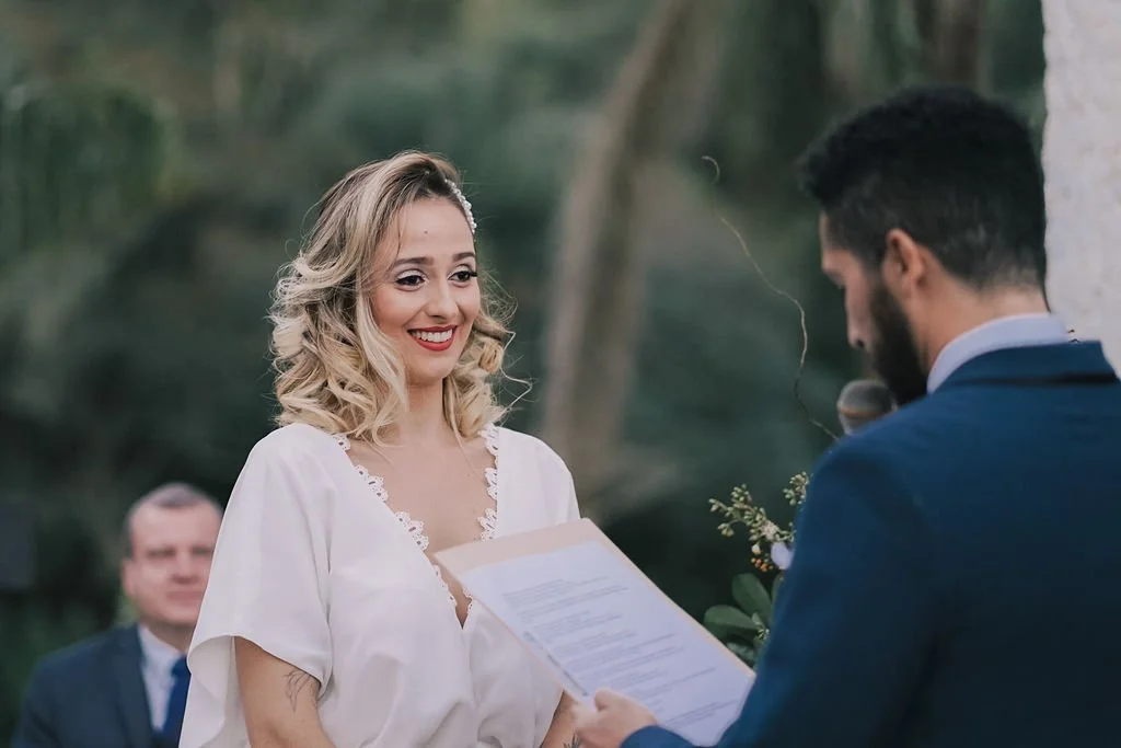 Casal em casamento ao ar livre, mulher sorridente, homem lendo votos.