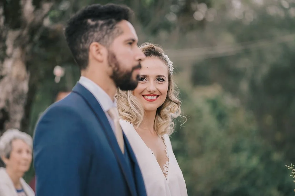 Homem e mulher em casamento ao ar livre, mulher sorrindo para a câmera, fundo com árvores e outra pessoa ao fundo.