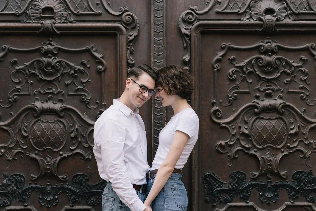 Um casal jovem com roupas casuais, sorrindo e ficando de cabeça próxima, em frente a uma porta de madeira com detalhes esculpidos.