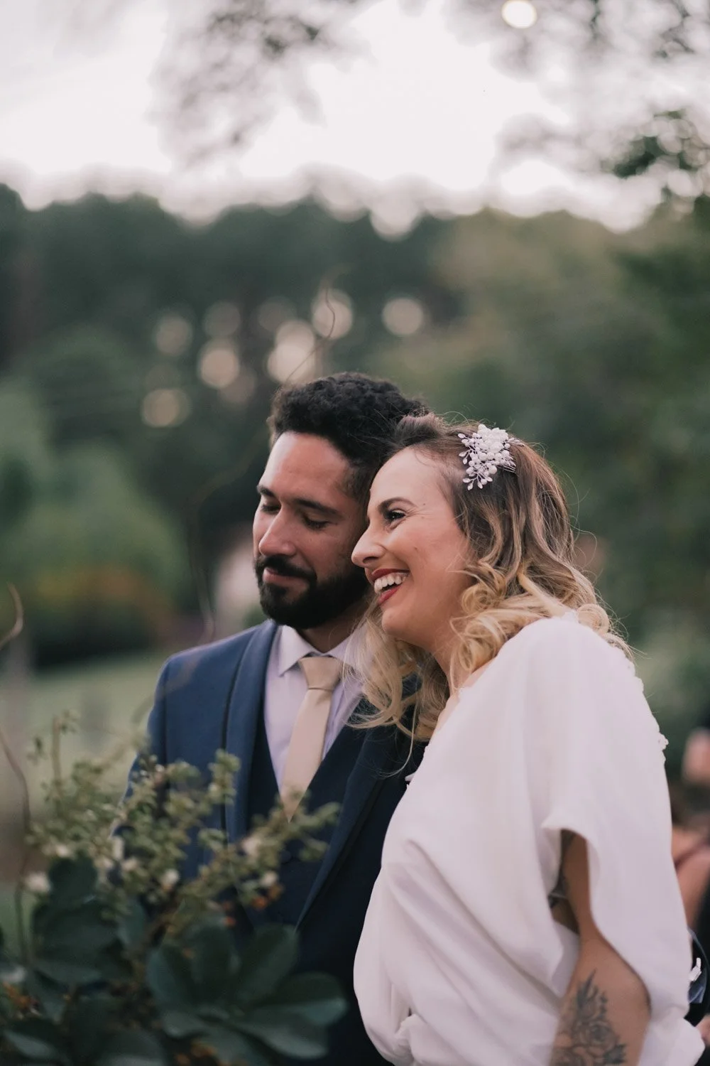 Casal sorrindo ao ar livre durante casamento, a mulher usa vestido branco e cabelo com acessório floral, o homem usa terno azul e gravata bege, em um ambiente com vegetação e árvores ao fundo.