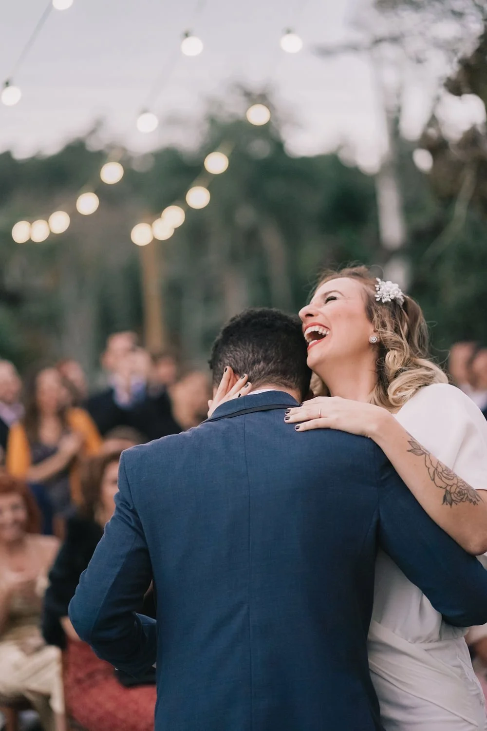 A couple dancing joyfully at a wedding, with string lights and guests in the background.