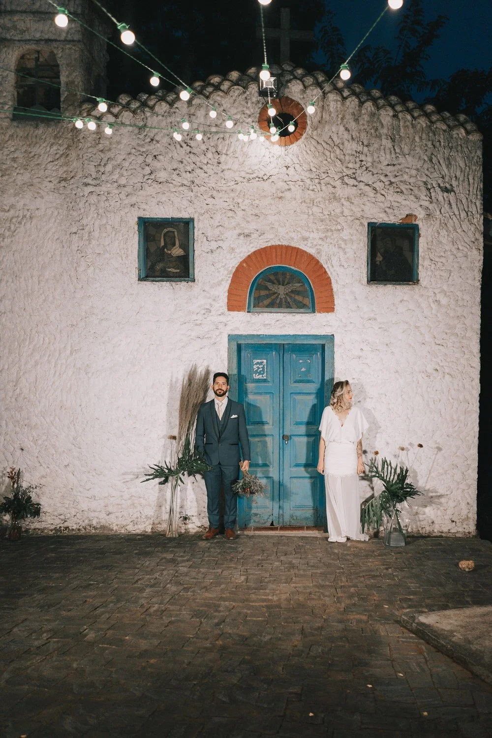 Casal de casamento posando em frente a uma parede de igreja antiga com portas azuis e janelas. A mulher veste vestido branco e o homem terno escuro. Iluminação de festa ao redor com luzes penduradas.