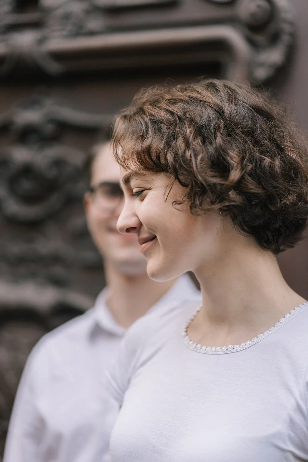 Duas mulheres sorrindo com cabelo cacheado, uma mais perto da câmera e a outra ao fundo, ao lado de uma parede decorada.