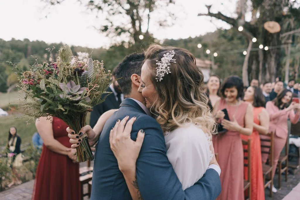 Casal de recém-casados abraçando-se durante uma cerimônia ao ar livre, com convidados ao fundo, árvores e luzes decorativas.