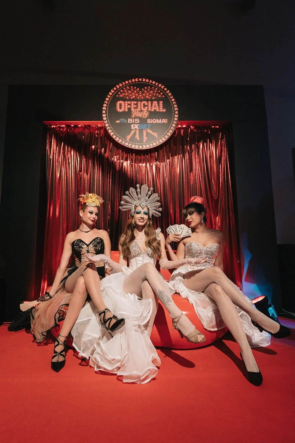 Três pessoas vestidas como bailarinas ou drag queens em roupas glamorosas, sentadas em um cenário de palco com cortina vermelha brilhante, uma delas segurando dinheiro, com um sinal de 'Official Party' acima.