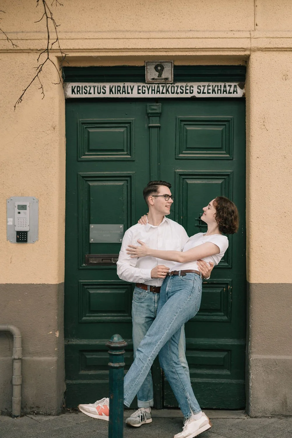 Casal feliz se abraçando na frente de uma porta verde, com uma placa em húngaro e um pequeno quadro de interfone ao lado, na rua.