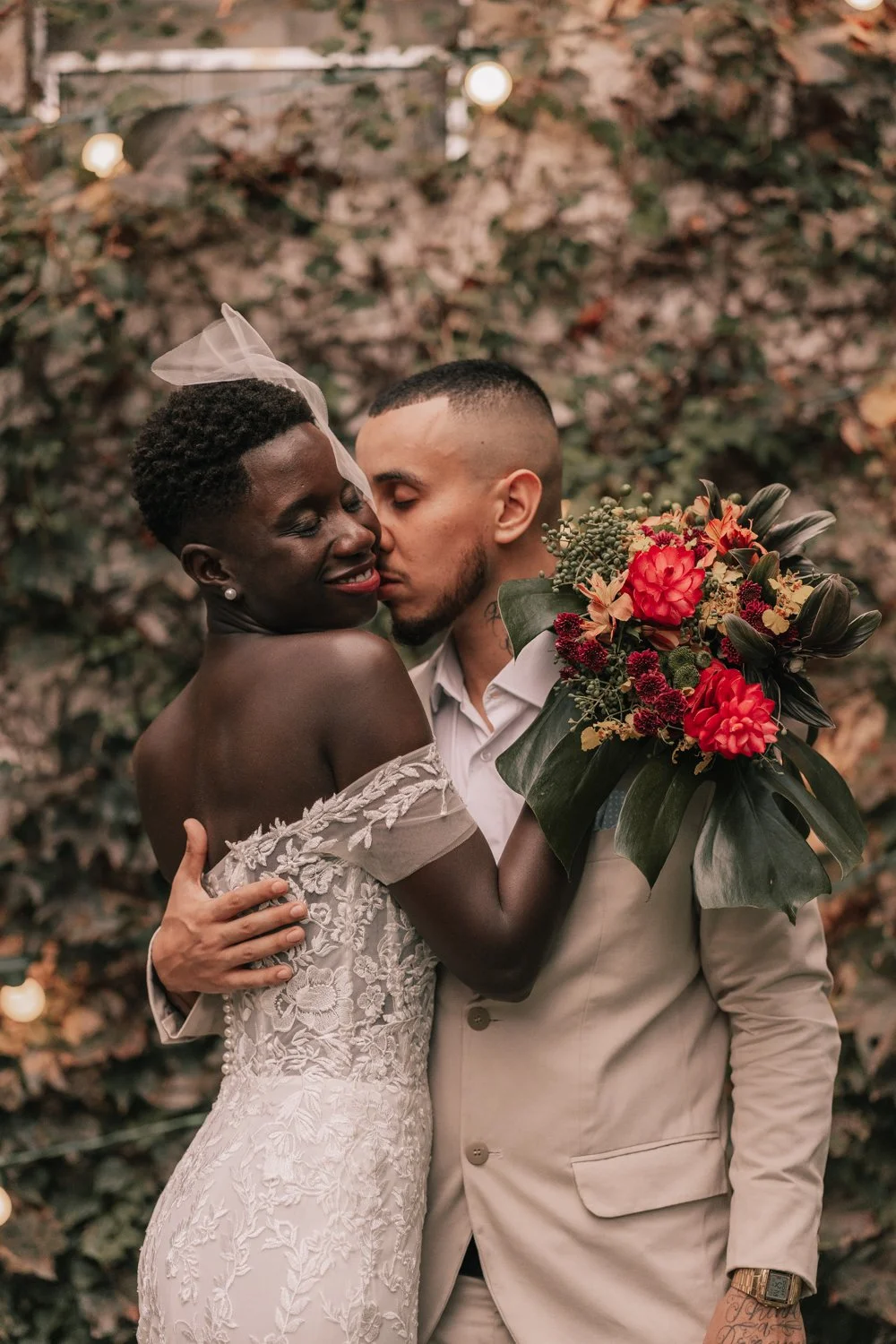 Casal apaixonado em um casamento, com o homem vestindo terno bege e a mulher em vestido de noiva com bordados, segurando um buquê de flores, enquanto se abraçam e se beijam, com fundo de foliagem.