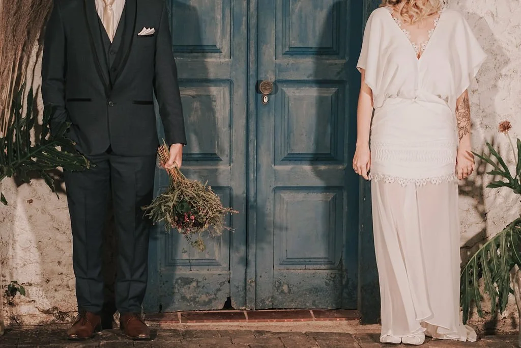 Pessoa vestida de terno preto e gravata, segurando um buquê de flores, ao lado de uma pessoa com vestido branco, ambos de frente para uma porta azul antiga.