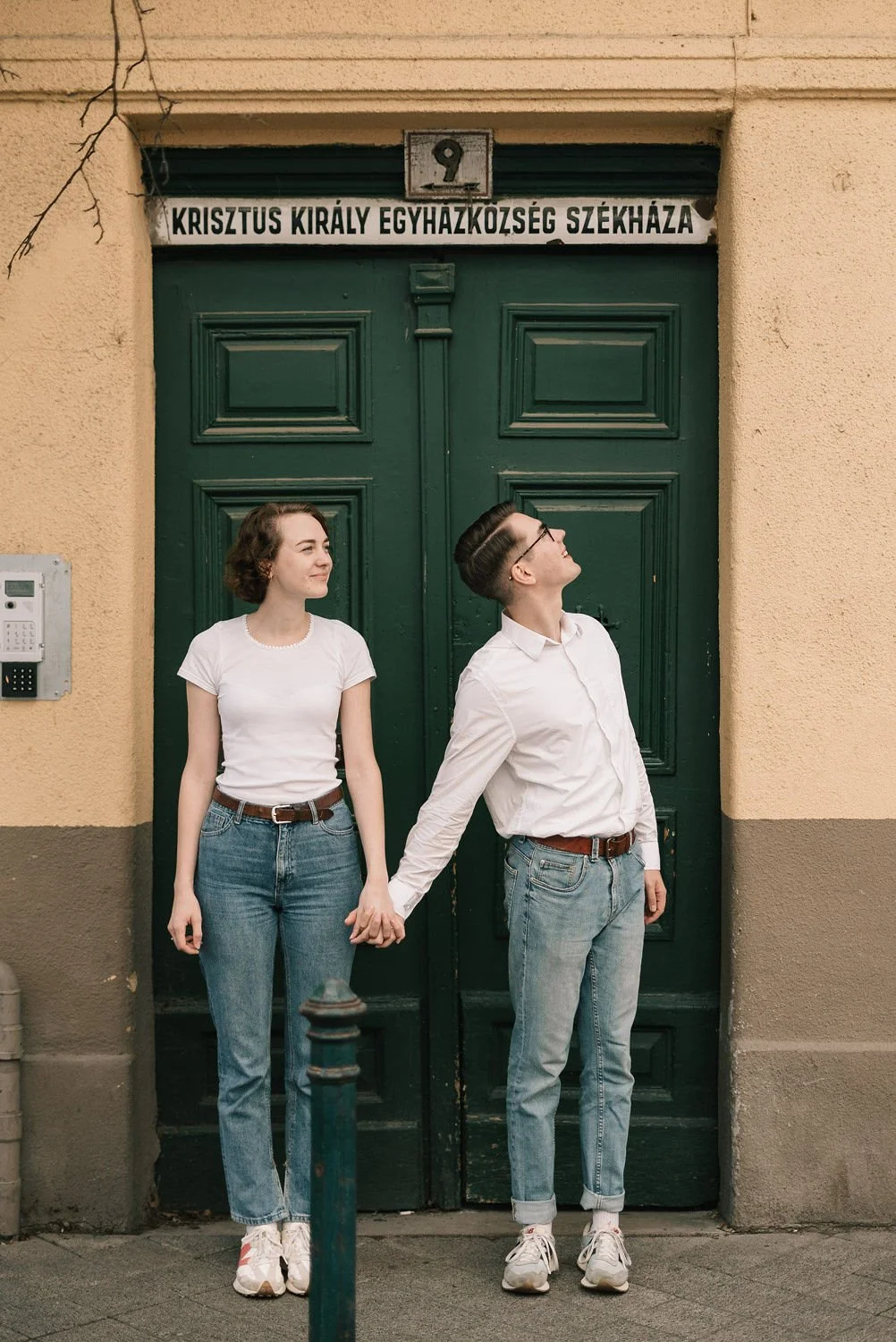 Jovens segurando as mãos na rua, posando em frente a uma porta verde com um sinal escrito em húngaro, usando roupas casuais e sorrindo, na cidade.