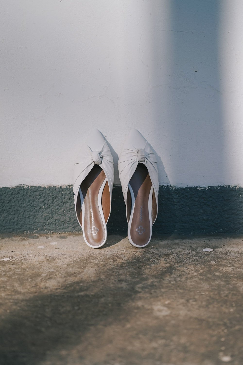 Sapatos femininos brancos com laços, apoiados contra uma parede branca